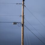 A solo crow or raven sits perched atop a telephone power pole with the sky and clouds in the background.